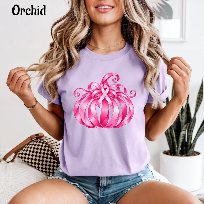 Op de afbeelding: Een lichtpaars t-shirt met een afbeelding van een pompoen gemaakt van roze linten. Het pompoenontwerp bevat een opvallend roze bewustzijnslint in het midden. Het woord Orchid is in zwarte letters in de linkerbovenhoek gedrukt.
