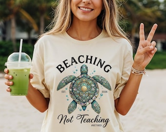 May include: A cream-colored t-shirt featuring a central graphic of a sea turtle with an intricate, colorful shell pattern. The text reads "BEACHING" above the turtle, "EST. 2026" on the sides, and "Not Teaching #RETIRED" in script below.