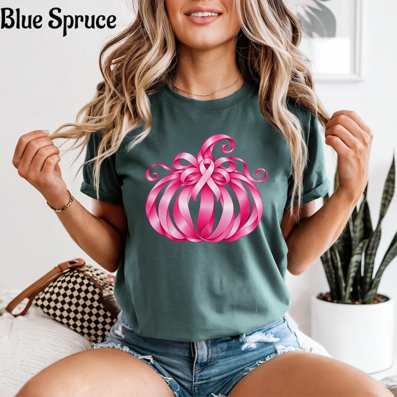 Op de afbeelding: Een donkergroen t-shirt met een afbeelding van een pompoen gemaakt van roze linten. Het ontwerp bevat een roze bewustwordingslint aan de bovenkant van de pompoen. De tekst Blue Spruce staat in de linkerbovenhoek van de afbeelding gedrukt.