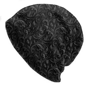 Op de afbeelding: Zwarte beanie-muts met een gedetailleerd, reliëf bloemmotief. Het ontwerp van de muts heeft een herhalend patroon van wervelende bladeren en wijnranken, waardoor een gestructureerd, driedimensionaal effect ontstaat. De muts is gemaakt van een zacht materiaal.