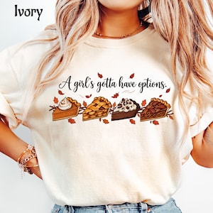 Puede incluir: Una camiseta de color marfil con el texto A girl's gotta have options. y un gráfico de cuatro porciones diferentes de tarta, incluyendo calabaza, manzana, chocolate y nuez pecana, rodeadas de pequeñas hojas de otoño en tonos naranja y marrón.