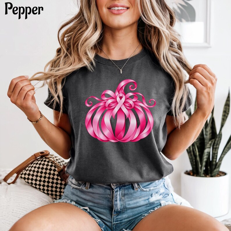 Op de afbeelding: Een donkergrijs t-shirt met korte mouwen en een afbeelding van een pompoen gemaakt van felroze bewustzijnslinten. Het woord Pepper is in het zwart in de linkerbovenhoek gedrukt. Het shirt heeft een ronde hals en een standaard pasvorm.