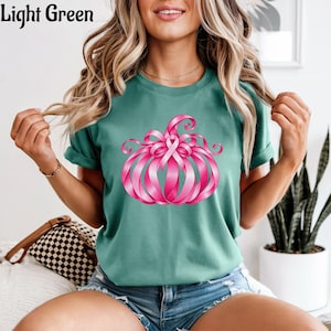 Op de afbeelding: Een lichtgroen T-shirt met korte mouwen en een afbeelding van een pompoen gemaakt van roze linten. Het ontwerp bevat een opvallend roze bewustzijnslint in het midden van de pompoen, met krullende lintsegmenten die de vorm vormen.