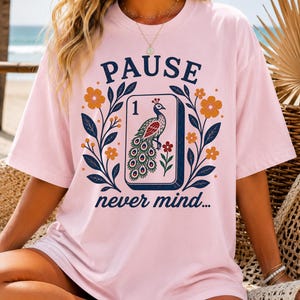 Op de afbeelding: Een lichtroze T-shirt met korte mouwen en een grafische print van een mahjongtegel met een pauwillustratie en het cijfer &eacute;&eacute;n. Het ontwerp bevat de tekst PAUSE en never mind, omringd door donkerblauwe bladtakken en oranje bloemen.