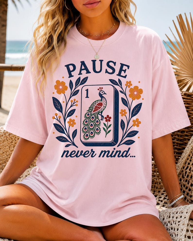 Op de afbeelding: Een lichtroze T-shirt met korte mouwen en een grafische print van een mahjongtegel met een pauwillustratie en het cijfer &eacute;&eacute;n. Het ontwerp bevat de tekst PAUSE en never mind, omringd door donkerblauwe bladtakken en oranje bloemen.