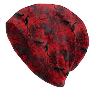 May include: A red beanie featuring a pattern of black crows perched on branches with red leaves. The hat has a soft, comfortable appearance, suitable for cooler weather. The design is repeated across the entire surface of the beanie.