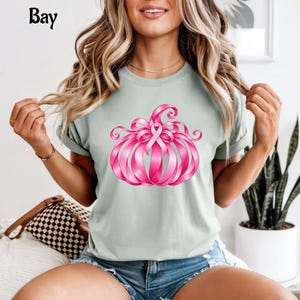 Op de afbeelding: Een lichtgroen T-shirt met korte mouwen en een afbeelding van een pompoen gemaakt van roze linten. Het ontwerp bevat een roze strik als symbool voor bewustwording boven op de pompoen. Het woord Bay staat in zwarte letters in de linkerbovenhoek.