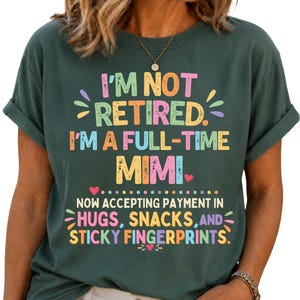 May include: A dark green short-sleeved t-shirt featuring the colourful text I'M NOT RETIRED. I'M A FULL-TIME MIMI. NOW ACCEPTING PAYMENT IN HUGS, SNACKS, AND STICKY FINGERPRINTS. The design includes small heart and petal accents in various pastel colours.