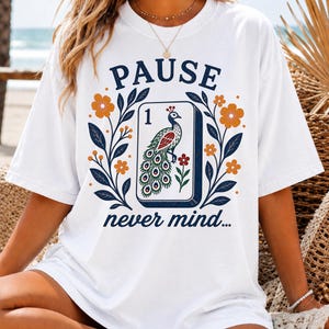 Op de afbeelding: Een wit t-shirt met een grafische print van een mahjongtegel met het cijfer &eacute;&eacute;n en een pauwillustratie. Het ontwerp bevat oranje bloemen, donkerblauwe bladeren en de tekst PAUSE erboven en never mind... eronder.