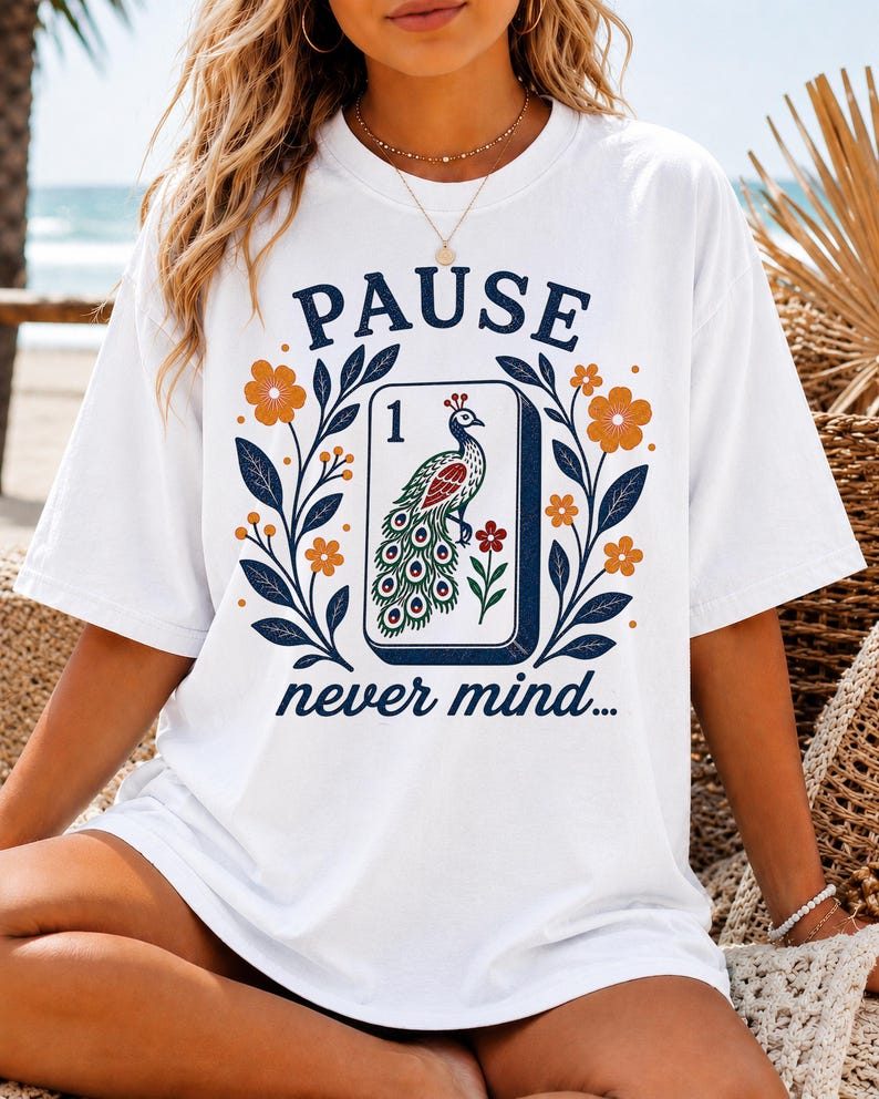 Op de afbeelding: Een wit t-shirt met een grafische print van een mahjongtegel met het cijfer &eacute;&eacute;n en een pauwillustratie. Het ontwerp bevat oranje bloemen, donkerblauwe bladeren en de tekst PAUSE erboven en never mind... eronder.