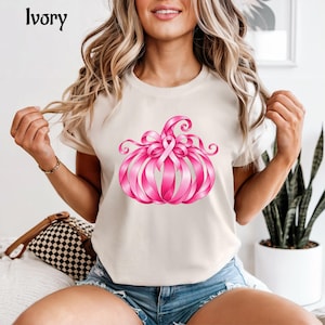 Op de afbeelding: Een ivoorkleurig T-shirt met korte mouwen en een afbeelding van een pompoen gemaakt van roze linten. Het woord Ivory staat in de linkerbovenhoek gedrukt. Het ontwerp bevat een centraal roze bewustzijnslint in de vorm van de pompoen.