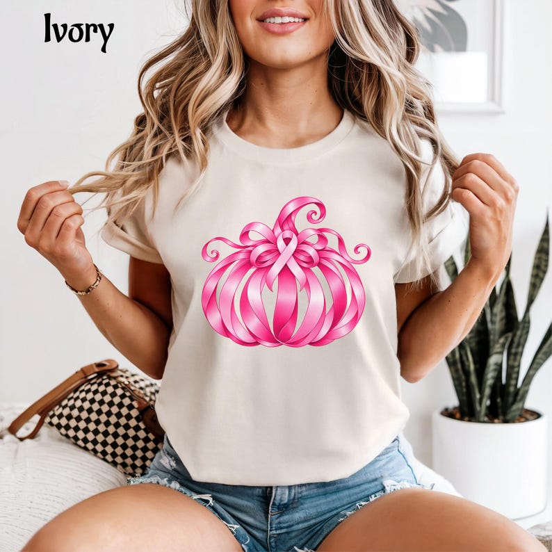 Op de afbeelding: Een ivoorkleurig T-shirt met korte mouwen en een afbeelding van een pompoen gemaakt van roze linten. Het woord Ivory staat in de linkerbovenhoek gedrukt. Het ontwerp bevat een centraal roze bewustzijnslint in de vorm van de pompoen.