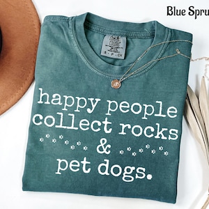 May include: A blue spruce coloured t-shirt with white text that reads happy people collect rocks & pet dogs. The shirt features a crew neckline and a Comfort Colors brand label on the inside collar. Small white paw print graphics are included.