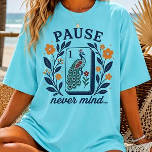 Op de afbeelding: Een lichtblauw t-shirt met korte mouwen en een grafische print van een mahjongtegel met een pauwillustratie, omringd door oranje en gele bloemen en donkerblauwe bladeren. De tekst PAUSE staat erboven en never mind... staat onder de tegel.