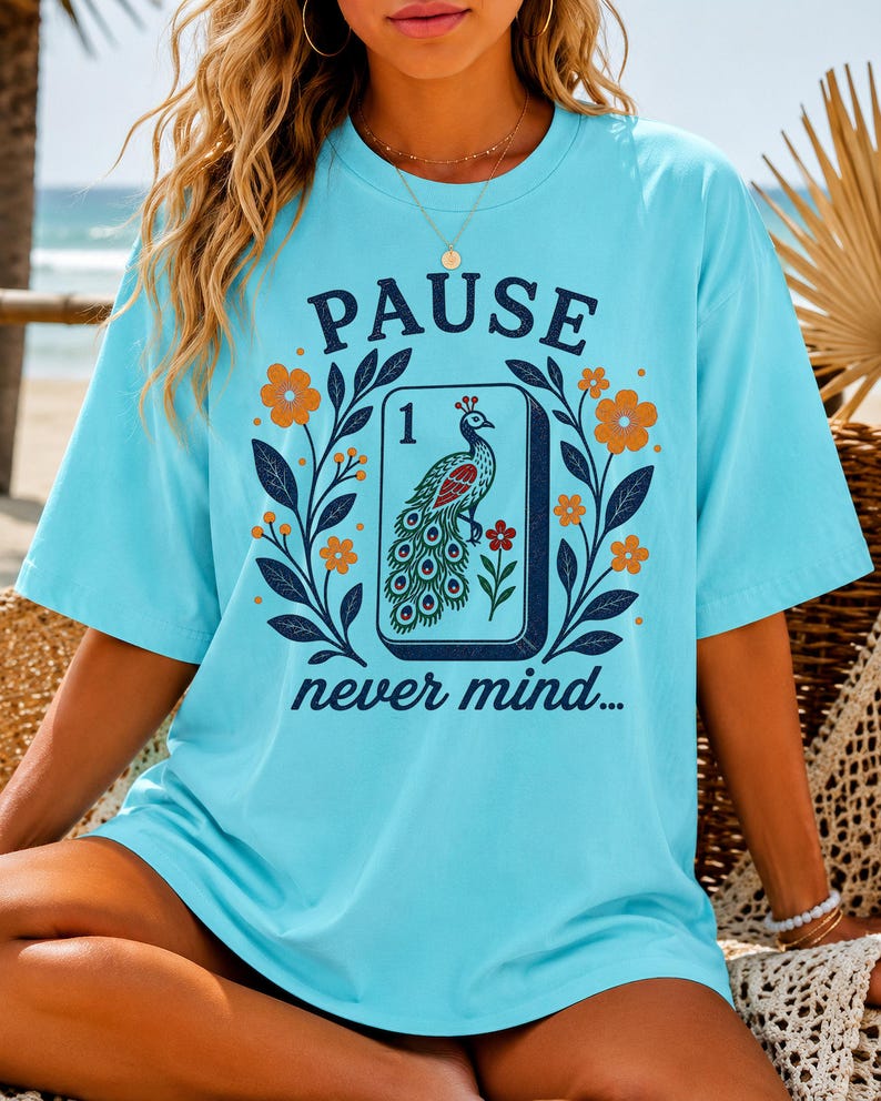 Op de afbeelding: Een lichtblauw t-shirt met korte mouwen en een grafische print van een mahjongtegel met een pauwillustratie, omringd door oranje en gele bloemen en donkerblauwe bladeren. De tekst PAUSE staat erboven en never mind... staat onder de tegel.