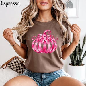 Op de afbeelding: Een bruin t-shirt met korte mouwen en een afbeelding van een pompoen gemaakt van roze linten. Het woord Espresso staat in het zwart in de linkerbovenhoek gedrukt. Het ontwerp bevat een centrale roze strik als symbool.