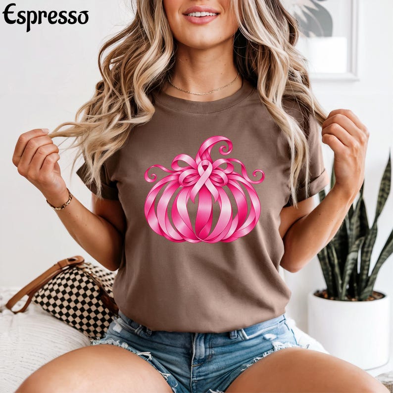 Op de afbeelding: Een bruin t-shirt met korte mouwen en een afbeelding van een pompoen gemaakt van roze linten. Het woord Espresso staat in het zwart in de linkerbovenhoek gedrukt. Het ontwerp bevat een centrale roze strik als symbool.