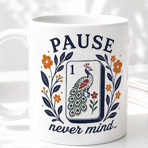 Op de afbeelding: Een witte keramische mok met een afbeelding van een mahjongtegel met het cijfer &eacute;&eacute;n en een pauw. Het ontwerp is omlijst door oranje bloemen en donkerblauwe bladeren, met de tekst PAUSE erboven en never mind... eronder in het blauw.