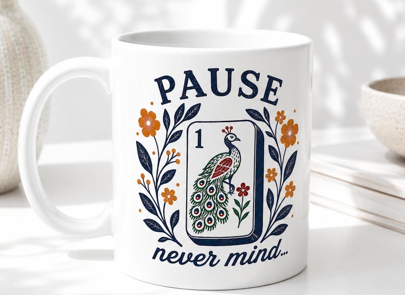 Op de afbeelding: Een witte keramische mok met een afbeelding van een mahjongtegel met het cijfer &eacute;&eacute;n en een pauw. Het ontwerp is omlijst door oranje bloemen en donkerblauwe bladeren, met de tekst PAUSE erboven en never mind... eronder in het blauw.
