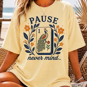 Op de afbeelding: Een lichtgeel t-shirt met korte mouwen en een grafische print van een mahjongtegel met een pauw en het cijfer &eacute;&eacute;n. De tekst PAUSE staat boven de tegel en never mind... staat eronder, omringd door bloemmotieven en blauwe bladeren.
