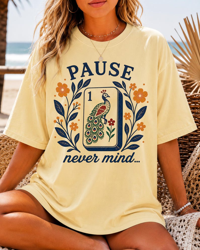 Op de afbeelding: Een lichtgeel t-shirt met korte mouwen en een grafische print van een mahjongtegel met een pauw en het cijfer &eacute;&eacute;n. De tekst PAUSE staat boven de tegel en never mind... staat eronder, omringd door bloemmotieven en blauwe bladeren.