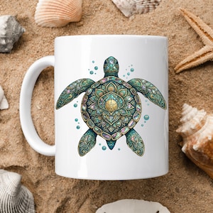 May include: White ceramic mug featuring a detailed, teal and gold sea turtle design. The turtle has an intricate mandala-style pattern on its shell and is surrounded by bubbles. The mug is set on a sandy surface with seashells.