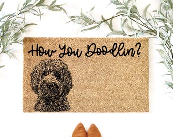 May include: A rectangular doormat with the text "How You Doodlin?" in black script. The mat features a detailed black and white illustration of a Goldendoodle dog. The mat is a light brown color and is surrounded by green foliage.