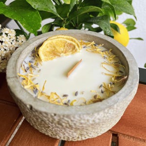 Könnte beinhalten: Ein runder, grauer Beton-Kerzenhalter mit einer weißen Kerze. Die Kerze ist mit getrockneten Zitronenscheiben, gelben Blütenblättern und Lavendelknospen verziert. Ein Holzdocht befindet sich in der Mitte. Die Kerze ist von grünen Blättern und einer Zitrone umgeben.