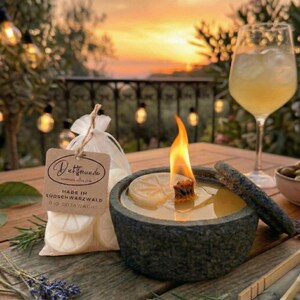 May include: A lit, stone candle holder with a lemon slice, next to a bag of white wax melts. The candle is burning with a bright flame. The background includes a cocktail, olives, and string lights.