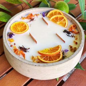 Könnte beinhalten: Runde, cremefarbene Kerze in einem hellgrauen Keramikbehälter. Verziert mit getrockneten Orangenscheiben, kleinen lila Blüten und anderen getrockneten Pflanzen. Die Kerze hat drei Holzdochte.