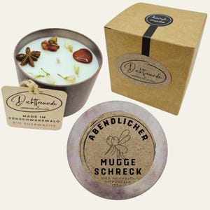 Könnte beinhalten: Eine handgemachte Sojawachskerze in einem braunen Metallbehälter, verziert mit getrockneten Blumen und Gewürzen. Die Kerze wird von einer Box und einem runden Behälter mit Text begleitet.