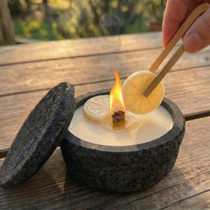May include: A lit candle in a dark gray stone bowl with a matching lid. A slice of lemon is being held over the flame with bamboo tongs. The candle has a white wax and a natural wick.