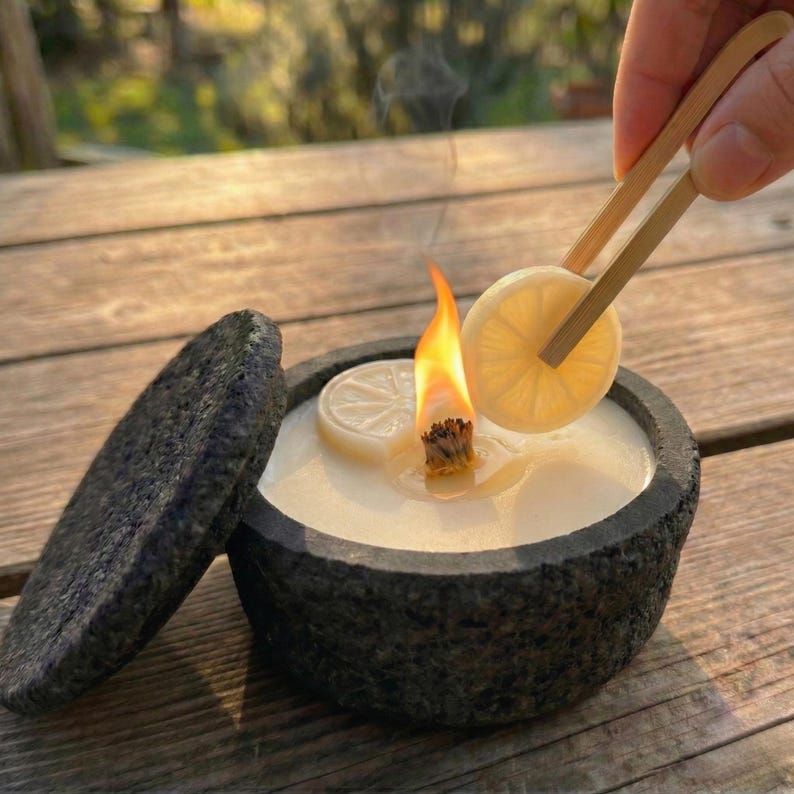 May include: A lit candle in a dark gray stone bowl with a matching lid. A slice of lemon is being held over the flame with bamboo tongs. The candle has a white wax and a natural wick.