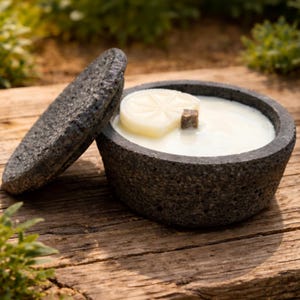 May include: A dark gray stone bowl with a lid, filled with white wax and a decorative candle. The candle has a citrus slice design and a small wooden cube. The bowl sits on a wooden surface.