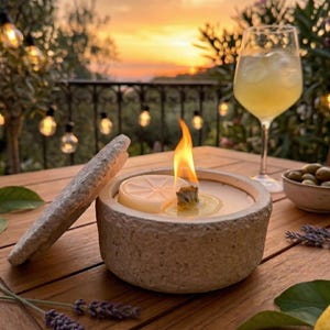May include: A lit citronella candle in a stone-like container with a lid, a glass of lemonade, and a small bowl of olives on a wooden table. The candle has a lemon slice and a flame. The background features a sunset and string lights.