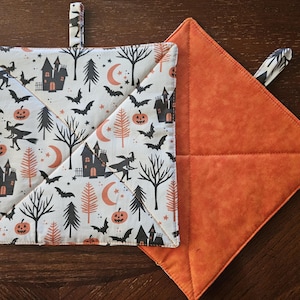 May include: Two square Halloween-themed pot holders. One features a white background with a repeating pattern of black haunted houses, bats, pumpkins, witches, and trees. The other is solid orange.