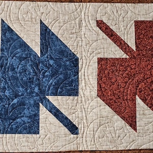 May include: A rectangular quilted table runner featuring four large leaf-shaped appliques in yellow, blue, red, and brown. The background fabric is a light cream color with a swirling stitched pattern that continues across the entire surface.