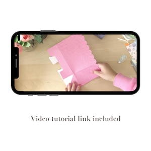 May include: A smartphone displays a crafting tutorial. The video shows a person folding a pink, polka-dotted paper with a scalloped edge. The text "Video tutorial link included" is at the bottom of the screen.