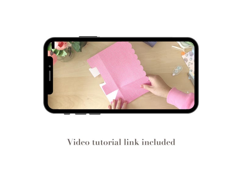 May include: A smartphone displays a crafting tutorial. The video shows a person folding a pink, polka-dotted paper with a scalloped edge. The text "Video tutorial link included" is at the bottom of the screen.