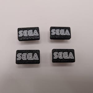 Puede incluir: Cuatro peque&ntilde;os parches rectangulares negros con el logotipo de SEGA en blanco. El texto tiene un aspecto texturizado. Los parches est&aacute;n dispuestos en forma cuadrada sobre un fondo blanco.