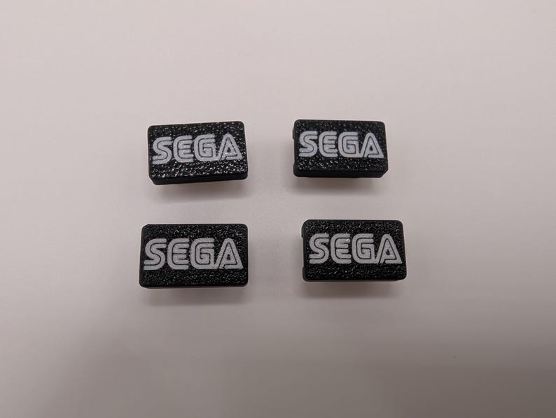 Puede incluir: Cuatro peque&ntilde;os parches rectangulares negros con el logotipo de SEGA en blanco. El texto tiene un aspecto texturizado. Los parches est&aacute;n dispuestos en forma cuadrada sobre un fondo blanco.