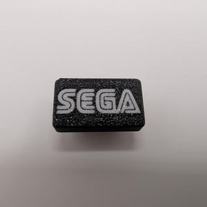 Puede incluir: Un objeto rectangular negro con la palabra "SEGA" en letras blancas. La superficie es texturizada, probablemente un bot&oacute;n o un emblema.