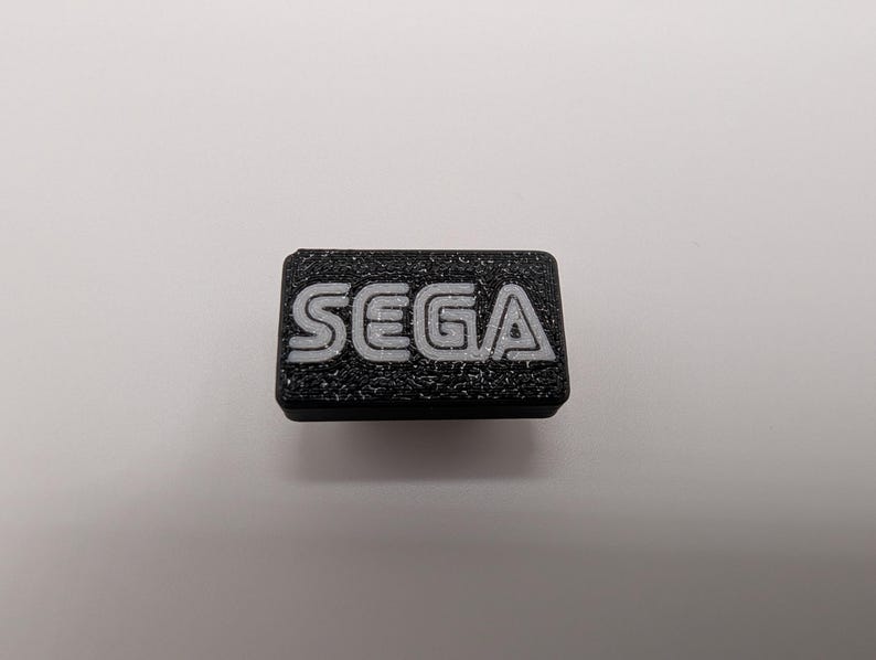 Puede incluir: Un objeto rectangular negro con la palabra "SEGA" en letras blancas. La superficie es texturizada, probablemente un bot&oacute;n o un emblema.