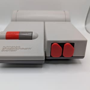 Puede incluir: Una consola gris del sistema de entretenimiento de Nintendo. La consola tiene un botón de encendido rojo y gris y dos puertos de controlador rojos. El texto "NINTENDO ENTERTAINMENT SYSTEM" está impreso en la parte delantera.