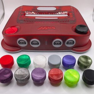 May include: A translucent red video game console featuring four controller ports and a top-loading cartridge slot. Below the console are twelve circular, textured buttons in various colours including red, green, white, grey, purple, olive, and black.