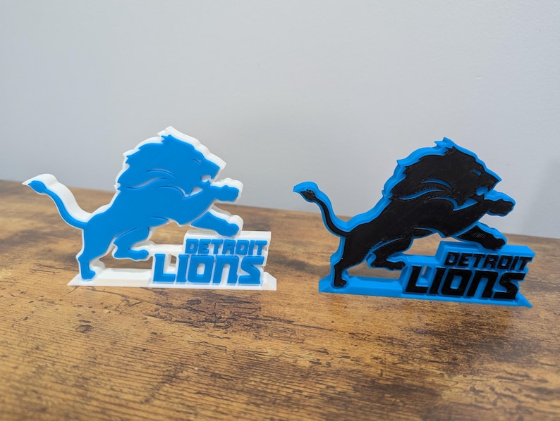 May include: Two 3D-printed Detroit Lions logos. One is light blue with a white base, the other is black with a light blue base. Both feature a leaping lion and the team name.