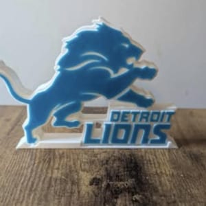 May include: A blue and white Detroit Lions logo, featuring a leaping lion, is displayed on a white base. The words "DETROIT LIONS" are printed in blue. The logo is set against a wooden surface.