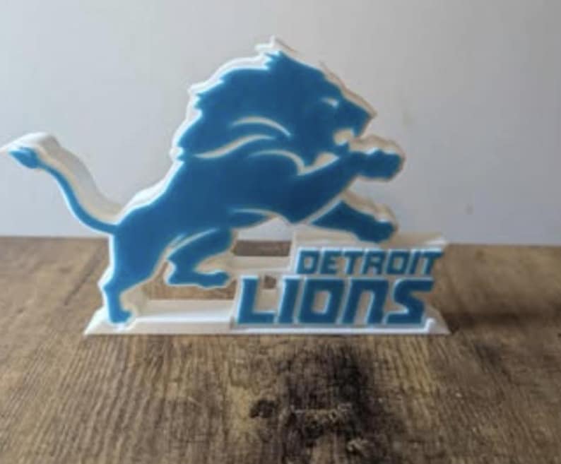 May include: A blue and white Detroit Lions logo, featuring a leaping lion, is displayed on a white base. The words "DETROIT LIONS" are printed in blue. The logo is set against a wooden surface.