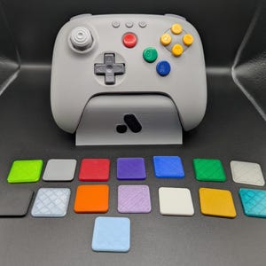 Puede incluir: Un mando de videojuegos gris con botones de colores y una cruceta, apoyado en un soporte a juego. Debajo hay varios botones cuadrados de colores, incluyendo verde, rojo y azul. El mando tiene un diseño retro.