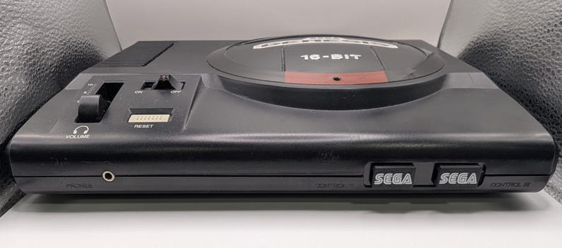 Puede incluir: Una consola Sega Genesis negra, una consola de videojuegos de 16 bits. La consola tiene un control de volumen, un bot&oacute;n de reinicio y dos puertos de controlador. El logotipo de Sega es visible en la parte frontal.
